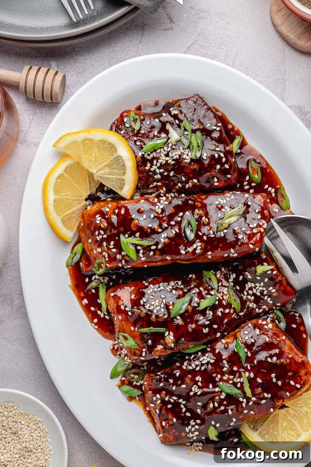 The final, finished Spicy Honey Garlic Salmon in a white serving dish with a thick, caramelized glaze. It is garnished with chopped fresh green onions, toasted sesame seeds, and small lemon wedges.