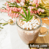 A white countertop with a cutting board topped with a clear mug full of cannabis hot cocoa with marshmallows, garnished with a cannabis leaf