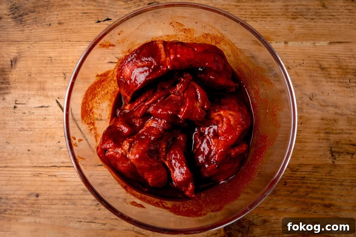 A glass bowl filled with raw char siu pork, coated in a red marinade. The bowl is on a wooden worktop.