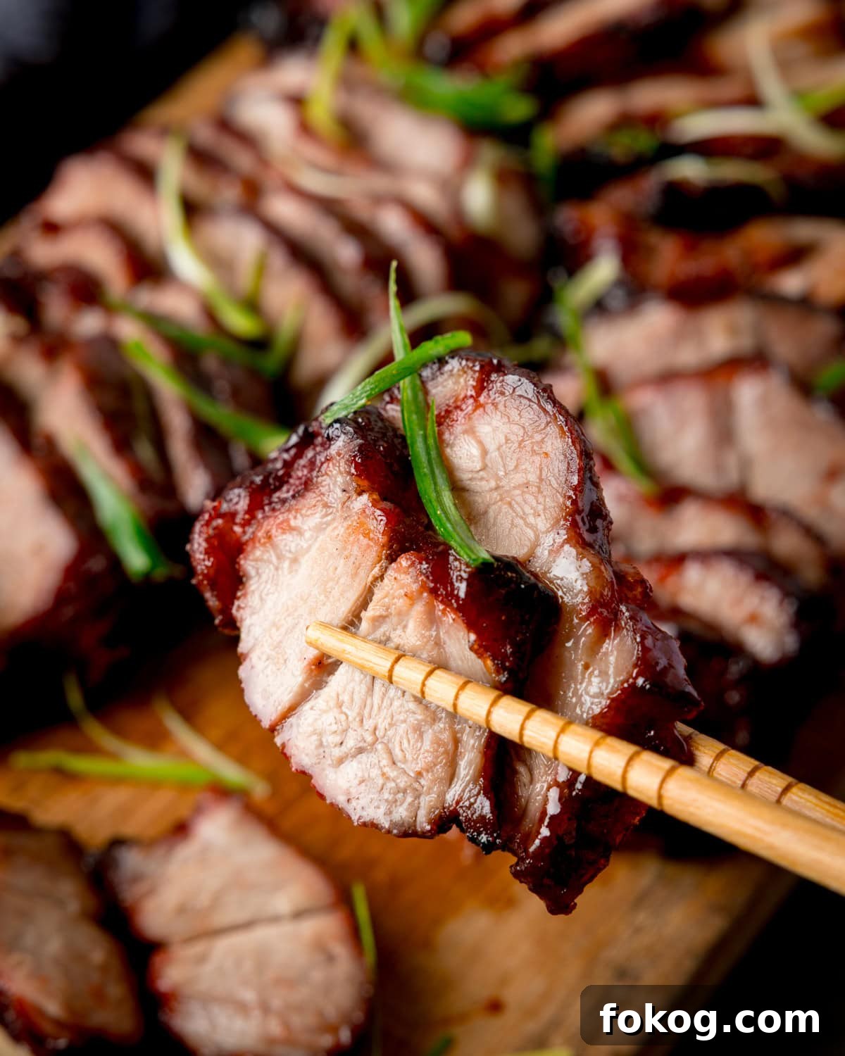 Chopsticks holding two slices of Chinese barbeque pork garnished with spring onions, more char siu on a wooden board in the background.