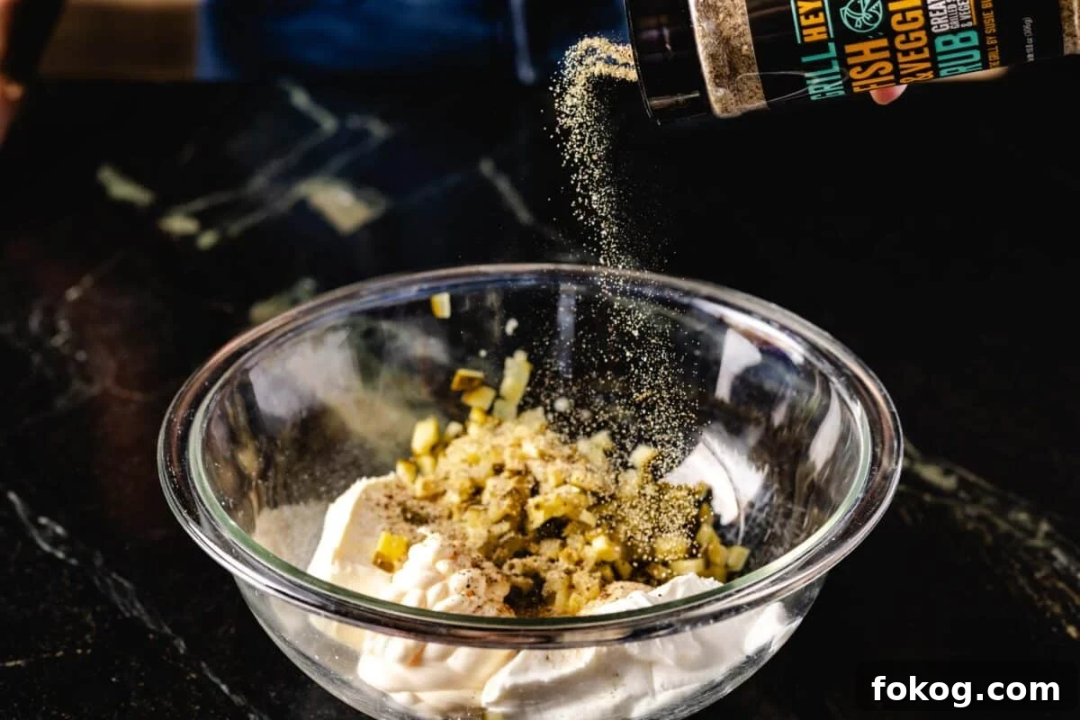 A bottle of rub being sprinkled into a clear bowl filled with mayo and diced pickles.