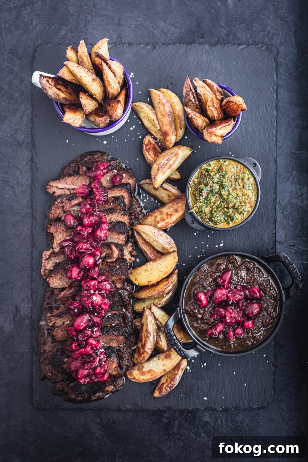 Cranberry-Braised Brisket: A Surprising Holiday Roast Delight 2 Thinly sliced brisket on a black slate with cranberry sauce on top, baked wedge potatoes and 2 little bowls of chimichurri and more cranberry sauce.