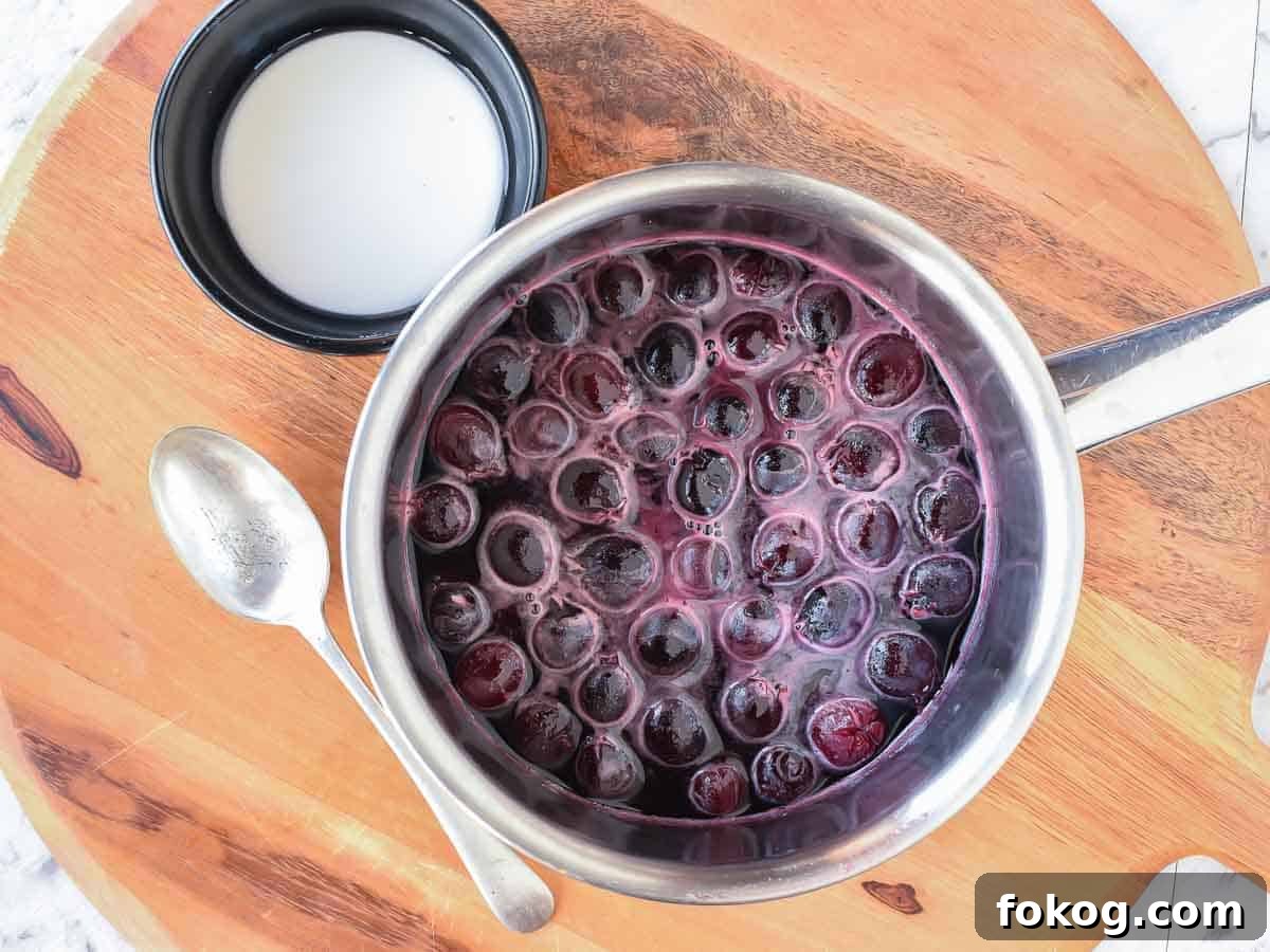Cherry Sauce using only 5 ingredients is easy to make at home. Perfect to use for cakes, cheesecakes, ice cream, pancakes, waffles or any dessert. #cherrysauce #easy #recipe #fresh#frozen #icecream #cheesecake #sweet #best #cherry #fruitsauce #dessert Cherries and liquid in a pot with white cornstarch slurry in black bowl and metal spoon beside viewed from above.
