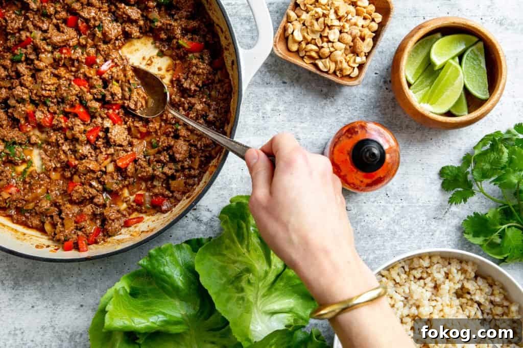 A hand spooning the beef lettuce wraps filling from a large skillet, with brown rice, lettuce leaves and peanuts alongside.