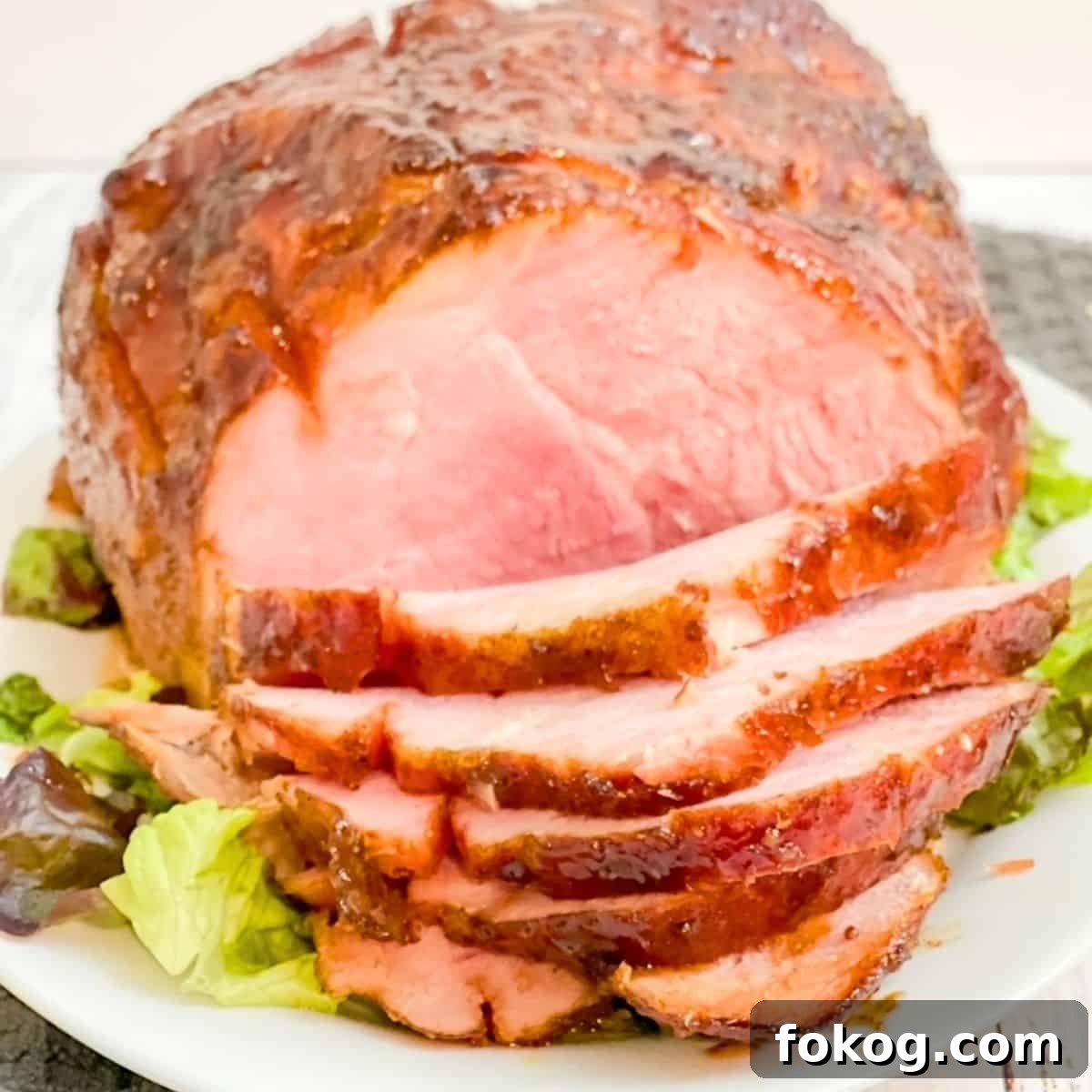 A twice smoked ham on a white serving platter, a few pieces sliced, sitting on top of green lettuce.