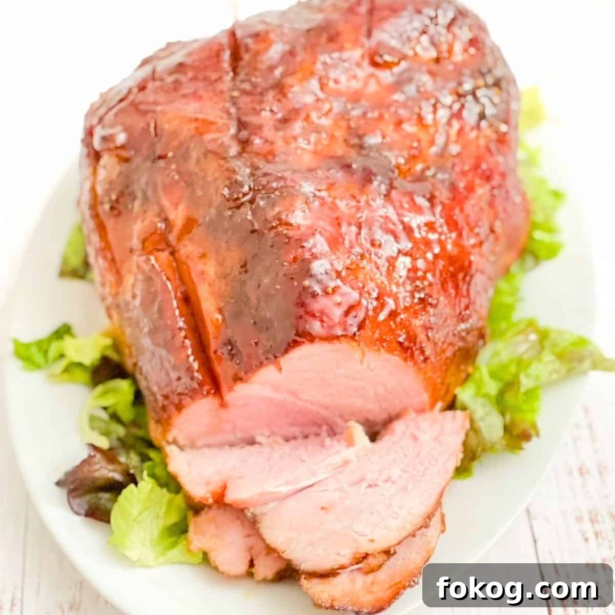 A double smoked ham on a white serving platter, a few pieces sliced, sitting on top of green lettuce.