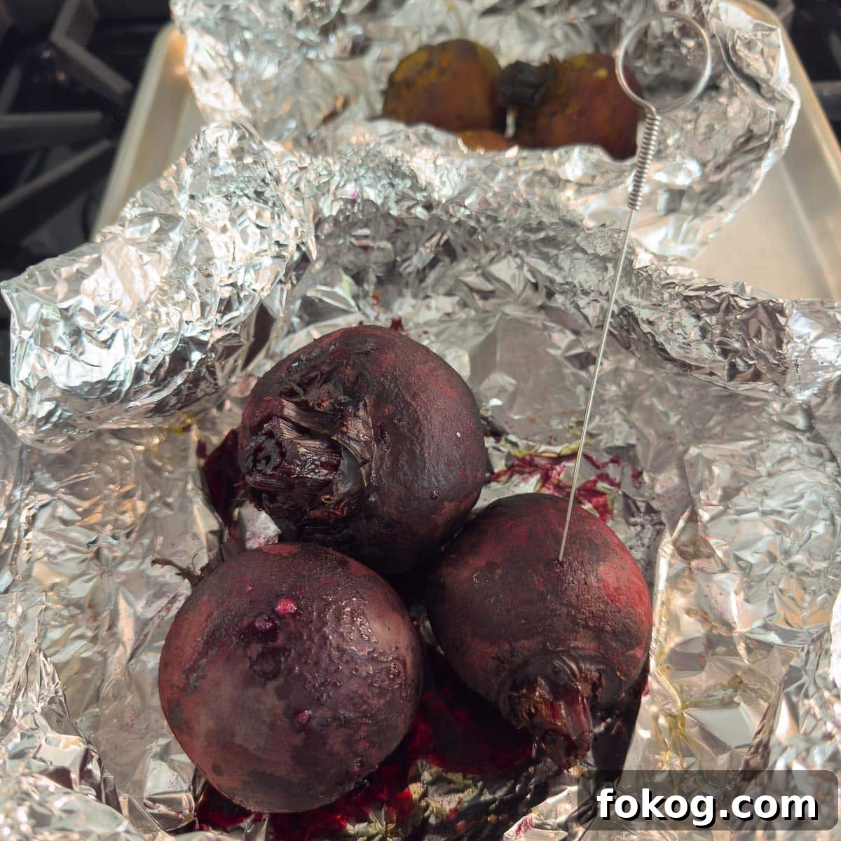 Roasted beets in foil with a thin metal skewer piercing one to show they are done.