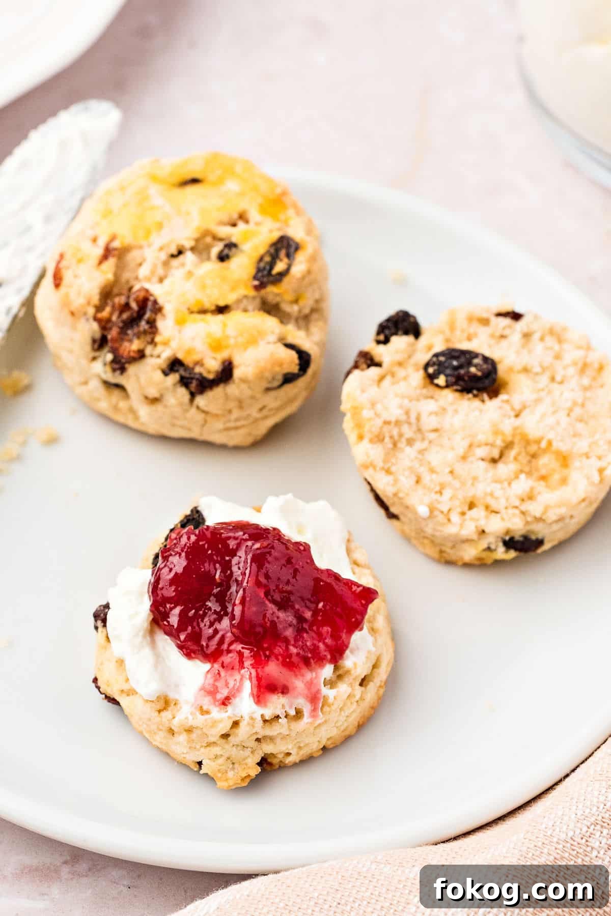Cream and jam topped scone with whole one on a white plate.