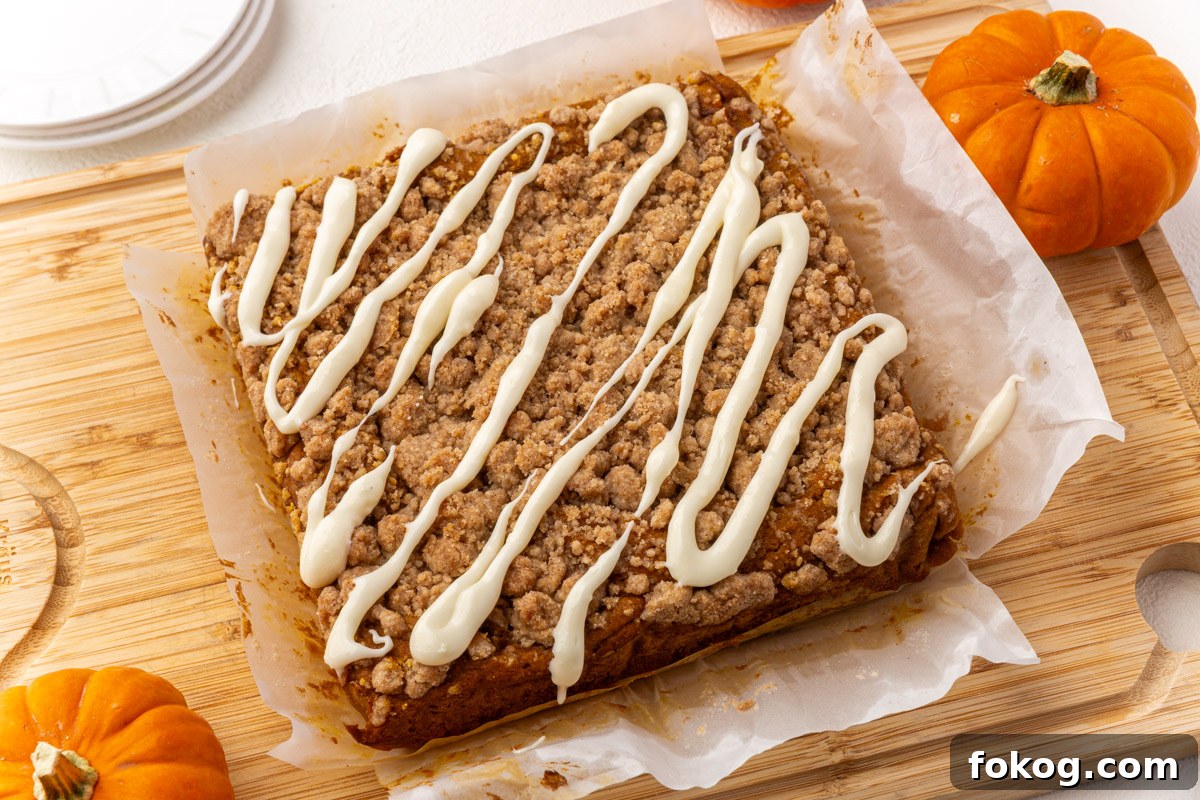 Freshly baked gluten-free pumpkin coffee cake with crumb topping and cream cheese glaze drizzle on parchment paper, surrounded by small pumpkins.
