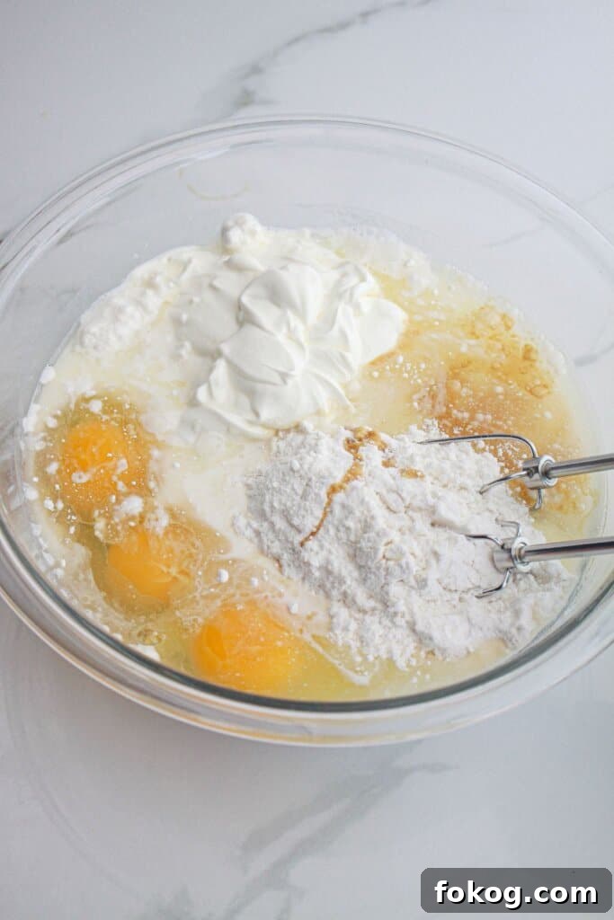 cake mix, sour cream, and eggs being mixed together with a hand mixer in a glass mixing bowl.