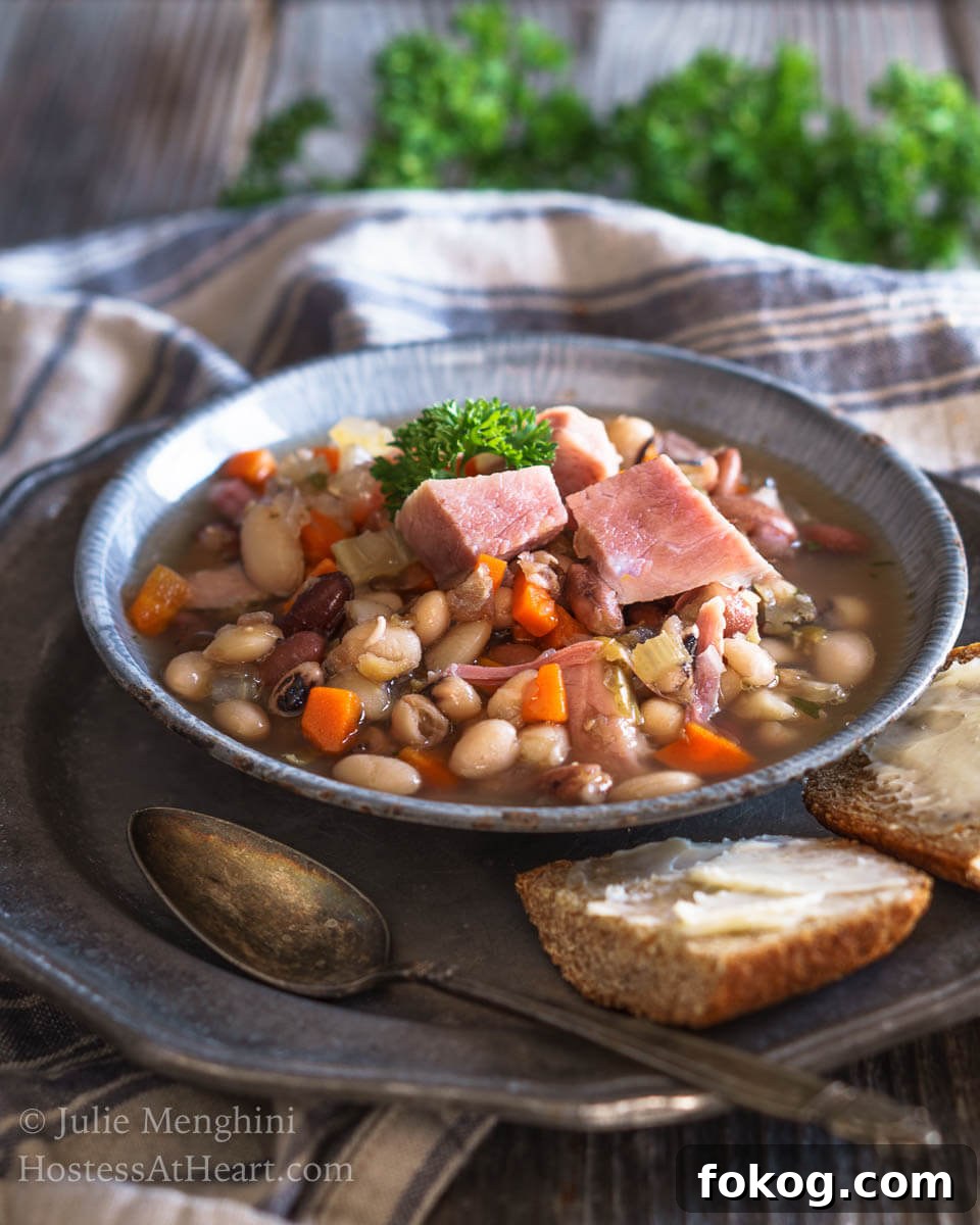 A bowl of Ham and Bean soup garnished with big chunks of ham. Sliced bread and a spoon sits next to the bowl on a metal plate.