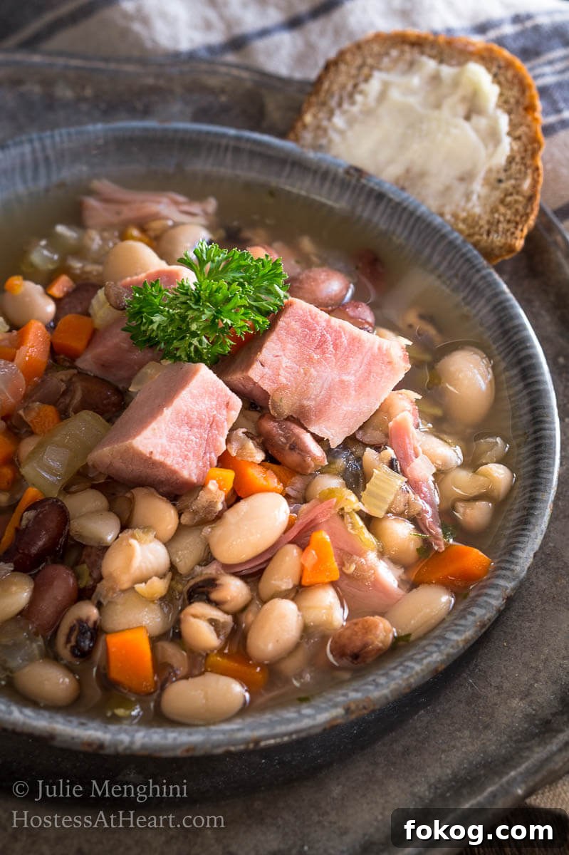 Top-down view of a bowl of Ham and Bean soup garnished with big chunks of ham. Sliced bread and a spoon sits next to the bowl on a metal plate.