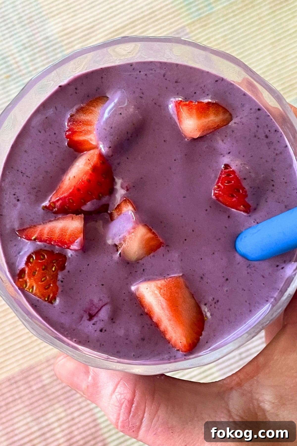 A blueberry banana and peanut butter smoothie with a blue straw held in a hand in a clear galss topped with some diced strawberries.