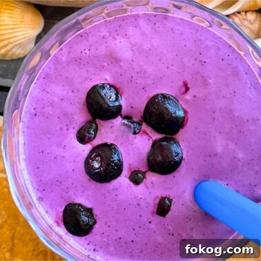A bluberry banana and peanut butter smoothie with blueberries on top on a tiled background with a blue straw and shells off to the side.